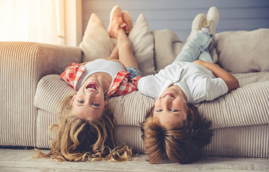 Natural Pharmaceuticals OmegaMarine Junior+ Naeratav tüdruk ja poiss lamavad tagurpidi diivanil, pea rippub allapoole ja jalad toetuvad vastu seljatuge.