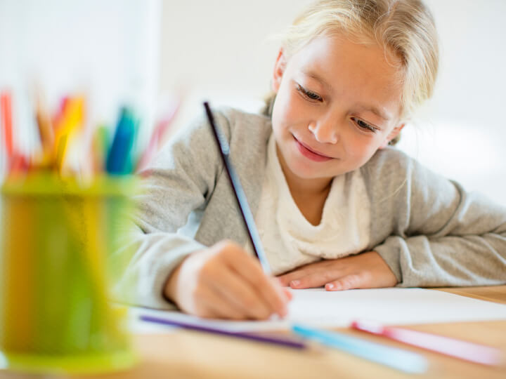Natural Pharmaceuticals OmegaMarine Junior+ Naeratav tüdruk kirjutab pliiatsiga laua taga, ees värvipliiatsid ja paber.