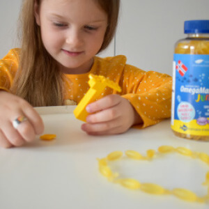 Natural Pharmaceuticals OmegaMarine Junior+ Tüdruk mängib kalakujulise marmelaadiga, mille kõrval on pudel oomega-3-vitamiini sisaldavaid lastevitamiine.