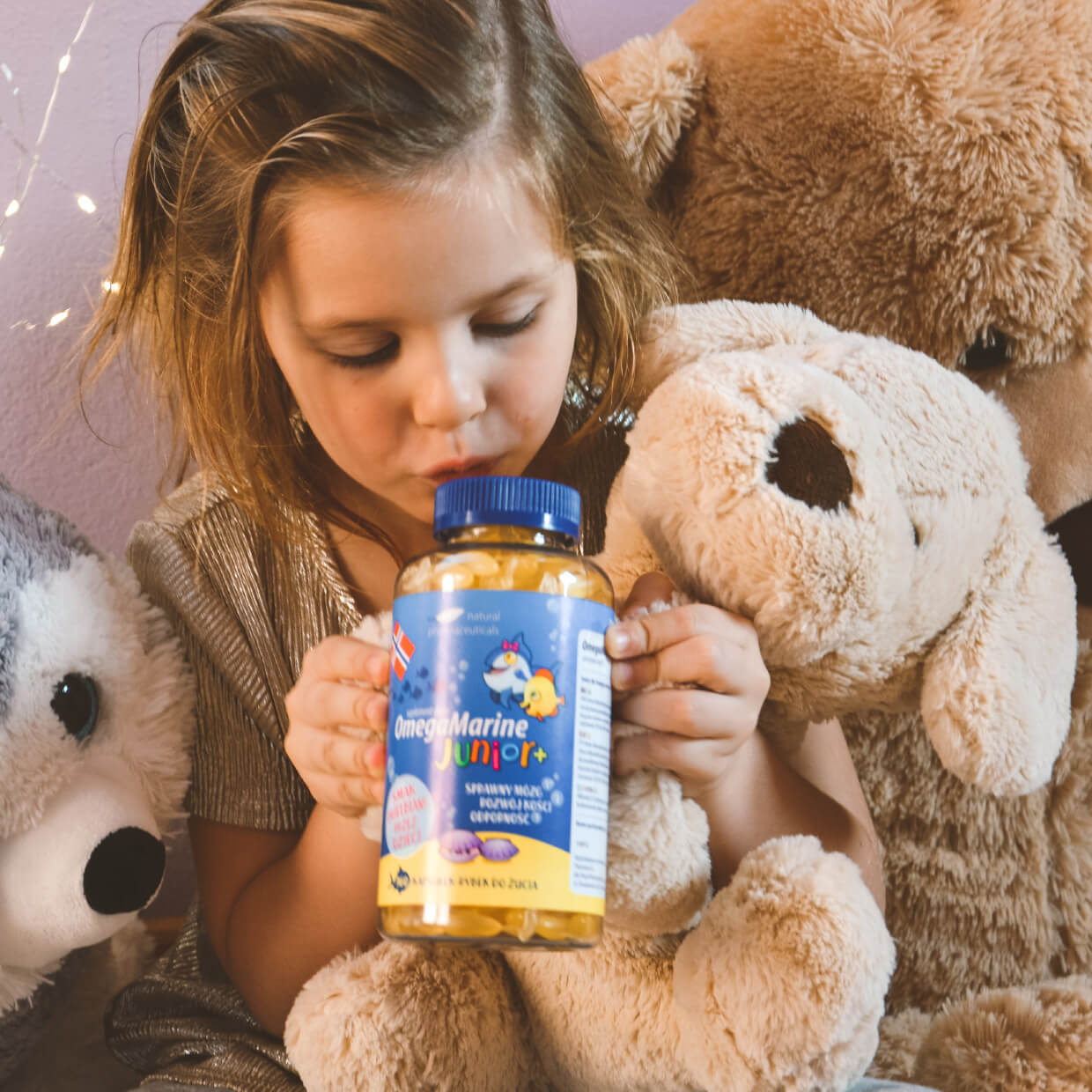 Natural Pharmaceuticals OmegaMarine Junior+ Tüdruk hoiab peos OmegaMarine Junior+ kalaõli vitamiine, ümbritsetud pehmete mänguloomadega turvalises keskkonnas.