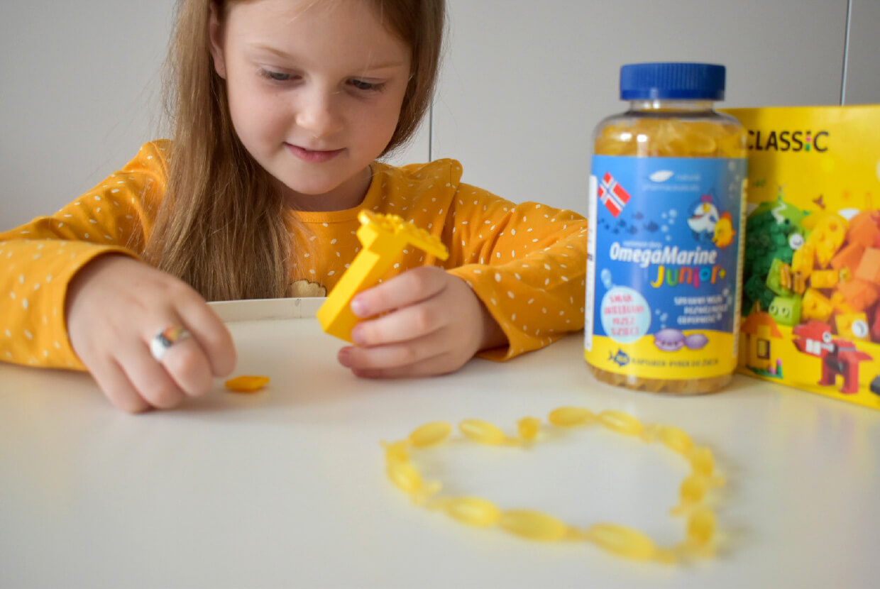 Natural Pharmaceuticals OmegaMarine Junior+ Tüdruk mängib kollaste Legodega laual, kus kõrval on OmegaMarine Junior+ kalaõli kapslitega pudel ja mänguklotsid.